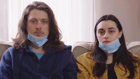 Couple sitting on a couch put on N95 masks and look into camera emotionless Stock Footage 129899081