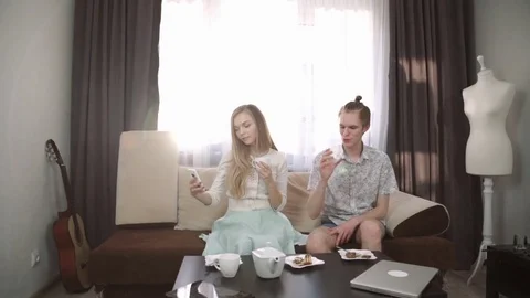 Couple is sitting on the couch sofa at home, Drink tea from a white tea set. Sun Stock Footage 74625034