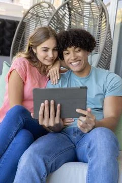 Couple sitting on cushioned sofa at covered patio, wearing T-shirts and jeans, Fotos Stock