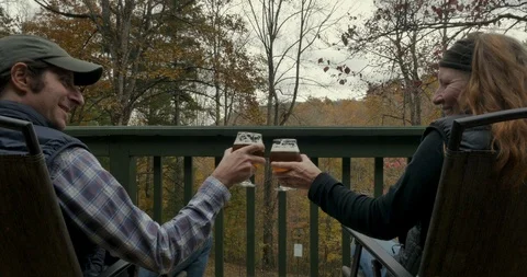 Couple sitting on a deck enjoying a view cheer with glasses of beer Stock Footage 112020156