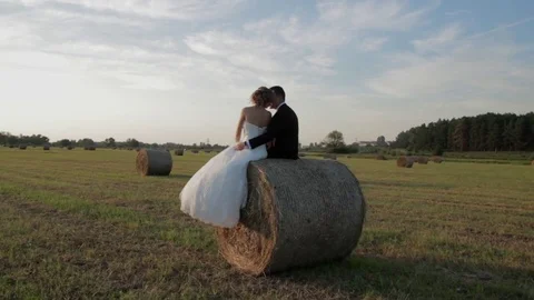 Couple sitting on a haystack. The bride and groom, sunset in the meadow Stock Footage 74714470