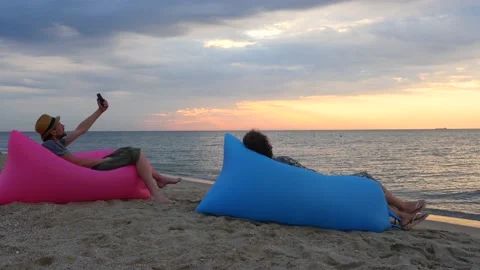 Couple sitting on inflatable sofa on beach at sunrise. Young man makes selfie Stock Footage 139979873