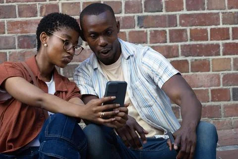 Couple sitting on low step by brick wall, leaning over checking smartphone, Fotos Stock