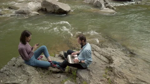 Couple sitting by mountain stream. Man working with laptop. Woman enjoying view Video stock 130356700