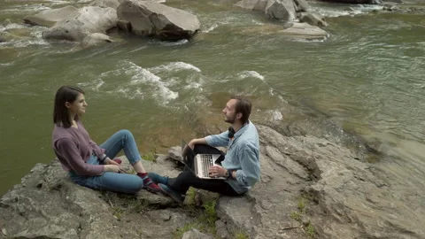 Couple sitting by mountain stream. Man taking photos of woman using smartphone Видео 130357274