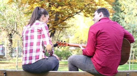Couple sitting on park bench, man playing guitar while woman singing Stock Footage 59556986
