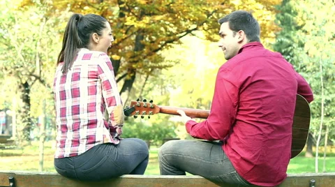 Couple sitting on park bench, man playing guitar while woman singing Stock Footage 59557003