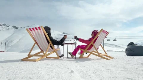 Couple sitting in pink striped deck chairs on snow toasting hot drinks in p.. Stock Footage 342962247