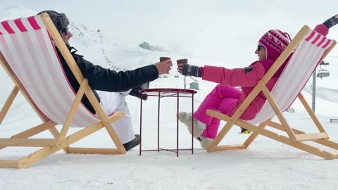 Couple sitting in pink striped deck chairs on snow toasting hot drinks in p.. Stock Footage 342962827