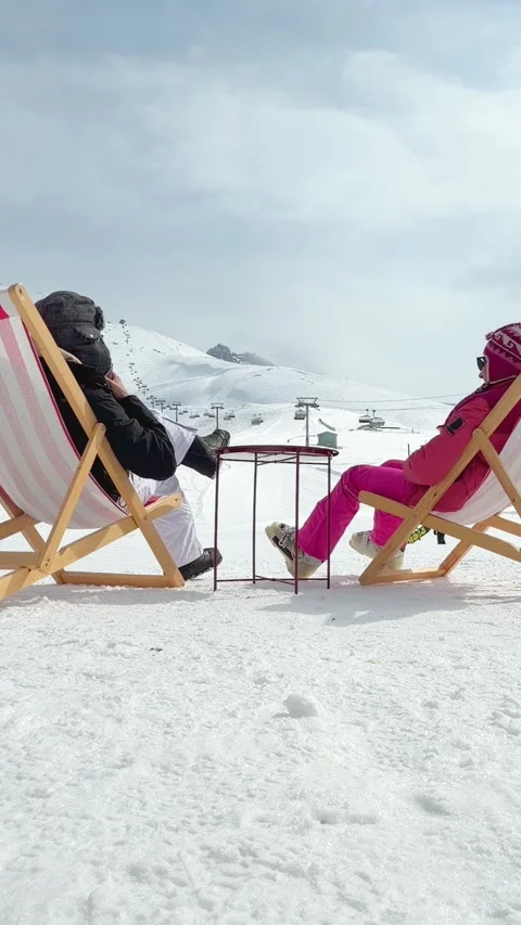 Couple sitting in pink striped deck chairs on snow toasting hot drinks in p.. Stock Footage 342962935