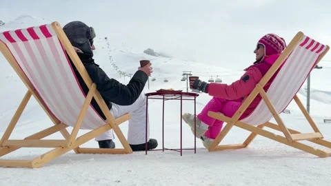 Couple sitting in pink striped deck chairs on snow toasting hot drinks in p.. Stock Footage 343556673