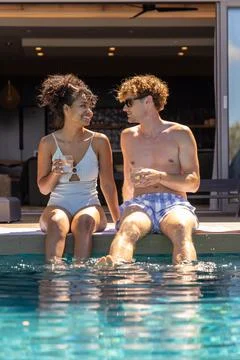 Couple sitting on pool deck edge dipping feet, holding clear glasses, wearing Fotos Stock