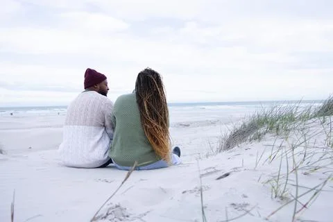 Couple sitting on sand dune at beach, wearing knit sweaters and beanie, showing Stock Photos