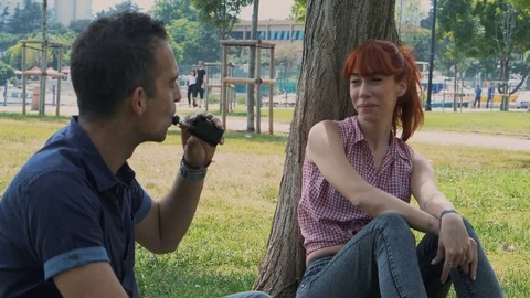 Couple smoking e-cigarette and relaxing in the park under a tree shadow Stock Footage 80934381