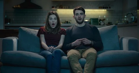 Couple on a sofa at home react to a shocking moment on tv Stock Footage 87636512