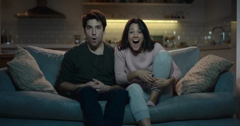Couple on a sofa react in amazement to something on the tv. Видео 87660490