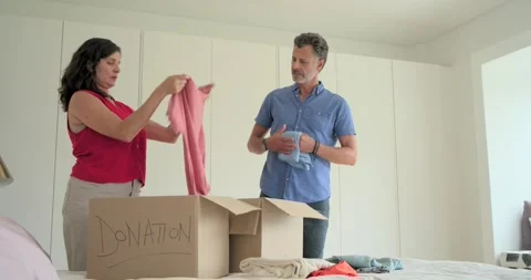 Couple sorting shirts on bed after closet cleanout, packing into box labeled Stock Footage 341852376