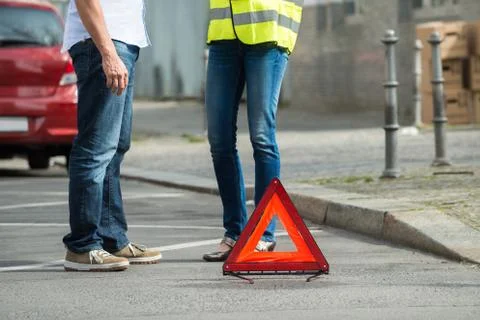 Couple Standing Near Triangular Warning Sign With Broken Down Car Foto stock
