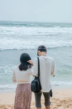 Couple Standing on Sandy Beach, Facing the Ocean Waves Under an Overcast Sk.. Stock Photos