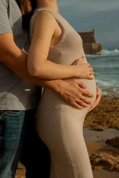 Couple stands together on sandy beach holding their baby bump during sunset n Stock Photos