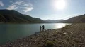 Couple strolling along bank of sunlit river holding hands, mountains on horizon 4k or 4k+ Resolution