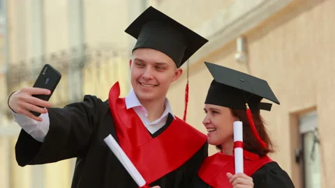 Couple of students in square caps and black red gowns makes a selfies Stock-Footage 191506077