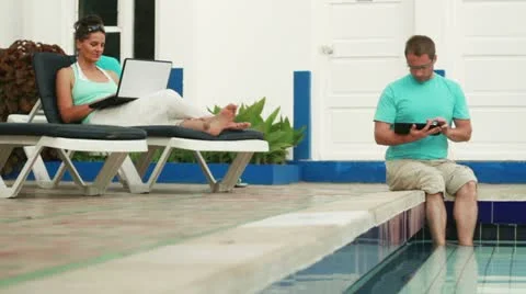 Couple with tablet computer and laptop relaxing on sunbeds by the swimming pool 스톡 동영상 10842101
