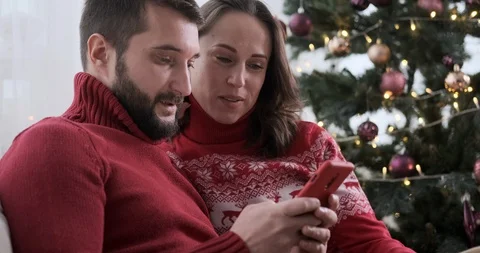 Couple using mobile phone next to Christmas tree at home Stock Footage 129424954