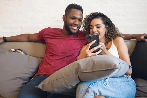 Couple using a mobile phone while sitting on a couch at home. Stock Photos