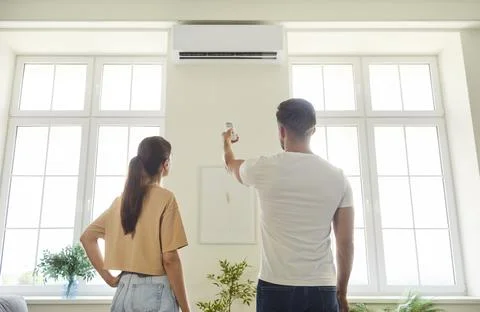 Couple using remote control to turn on air conditioner on wall of room at home Stockfoto's