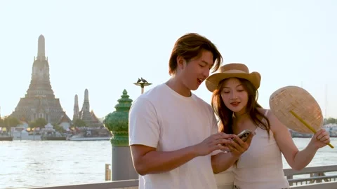 Couple Using Smartphone by Riverside Temple at Sunset in Bangkok Stock Footage 323434390