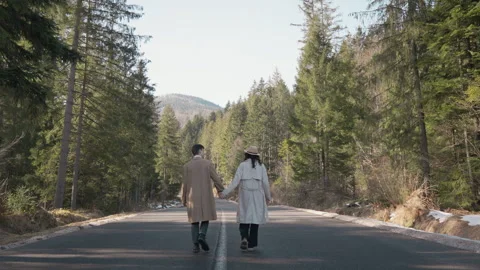 Couple walking hand in hand down a scenic forest road, surrounded by tall pine Stock Footage 340926489