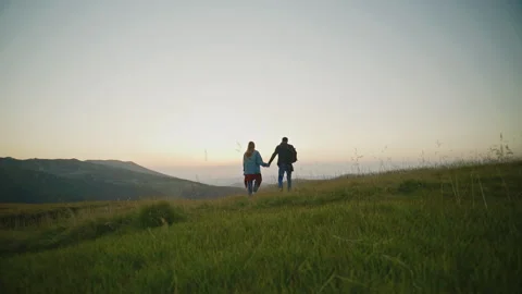 Couple walks hand in hand on a grassy hill during sunset near mountains Stock Footage 345683028