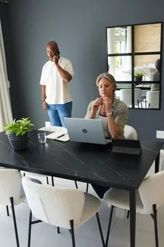 Couple working at home dining table, woman in blouse using laptop, man in white Stock Photos