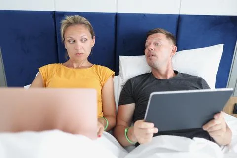 Couple working using gadgets in bed, man prying about woman Stock Photos