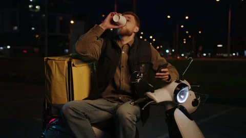 Courier drinks coffee sitting on a scooter and checks orders on his phone Stock Footage 214876476