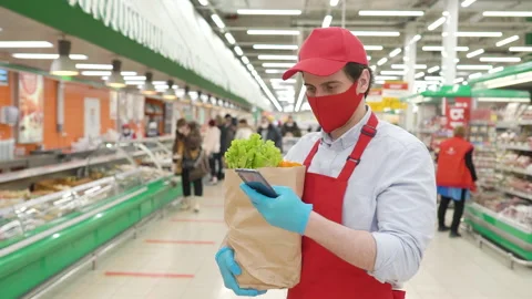 Courier in face mask, gloves and red uniform standing in supermarket with Stock Footage 148108564