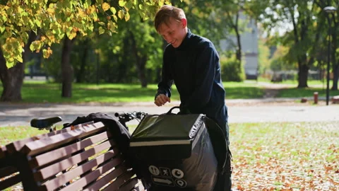 Courier on park bench, Insulated bag delivery in park, Courier places insulated Stock Footage 329575149
