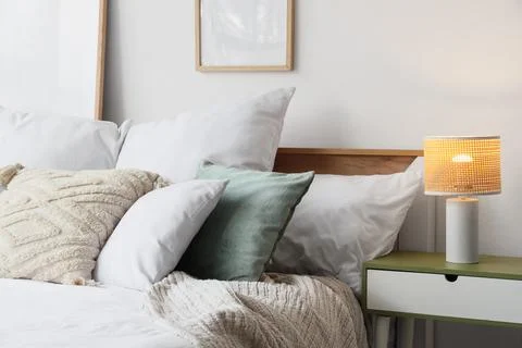 Cozy bed with soft pillows and glowing lamp on bedside table in bedroom Stock Photos