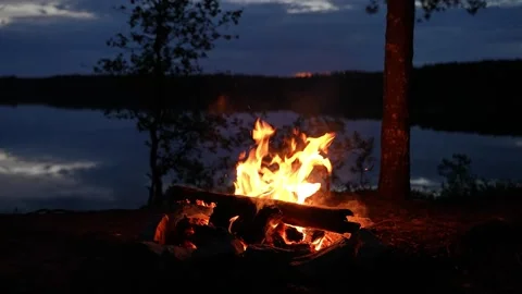 Cozy campfire burning on the shore of a tranquil lake at dusk, with glowing Video stock 312804103