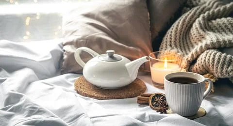 Cozy composition with a cup of tea, a teapot and a candle in the bed. Stock Photos