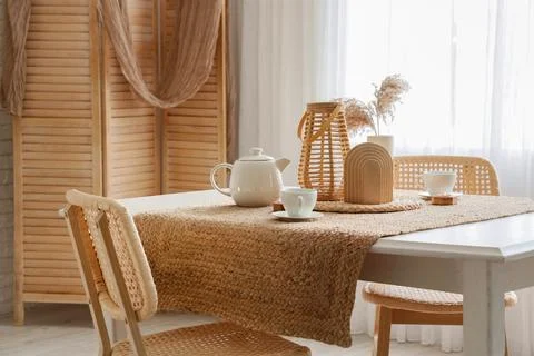 Cozy dining table setting with woven jute runner, teapot, cups, and rattan ch Stock Photos