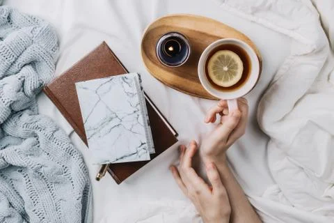 Cozy flatlay of woman's hands in bed with knitted sweater and books holding c Stock Photos