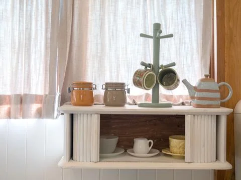 Cozy kitchen shelf with ceramic cups, teapot, and jars by soft curtain wind.. 스톡 사진
