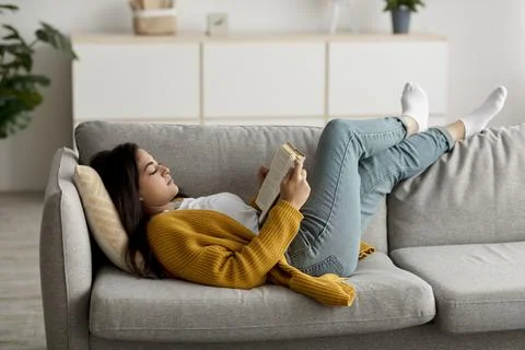 Cozy stay at home activities. Young arab woman reading her favorite book, lying Stock Photos