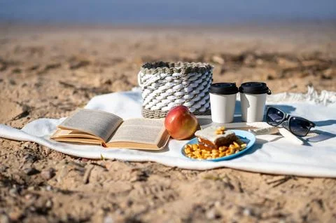 Cozy summer picnic setup on a sandy lake shore with coffee cups, snacks, a book, Stock Photos