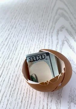 A cracked eggshell reveals a rolled-up banknote inside, placed on a light wooden Stock Photos