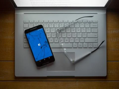 A cracked phone with the Facebook app loading sitting on a laptop. Stock Photos