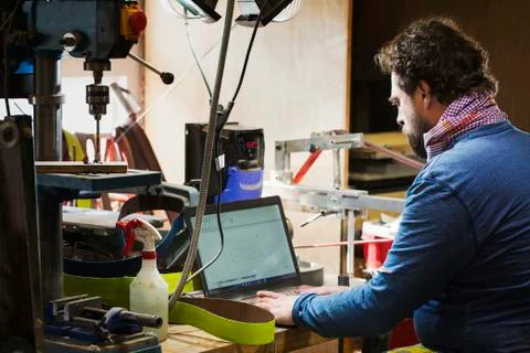 A craftsman at a desk in a workshop using a laptop computer on a cluttered desk. Stock Photos