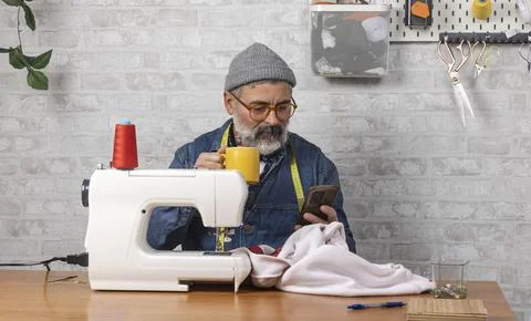 Craftsman drinking coffee and using smartphone while sewing Stock-Fotos
